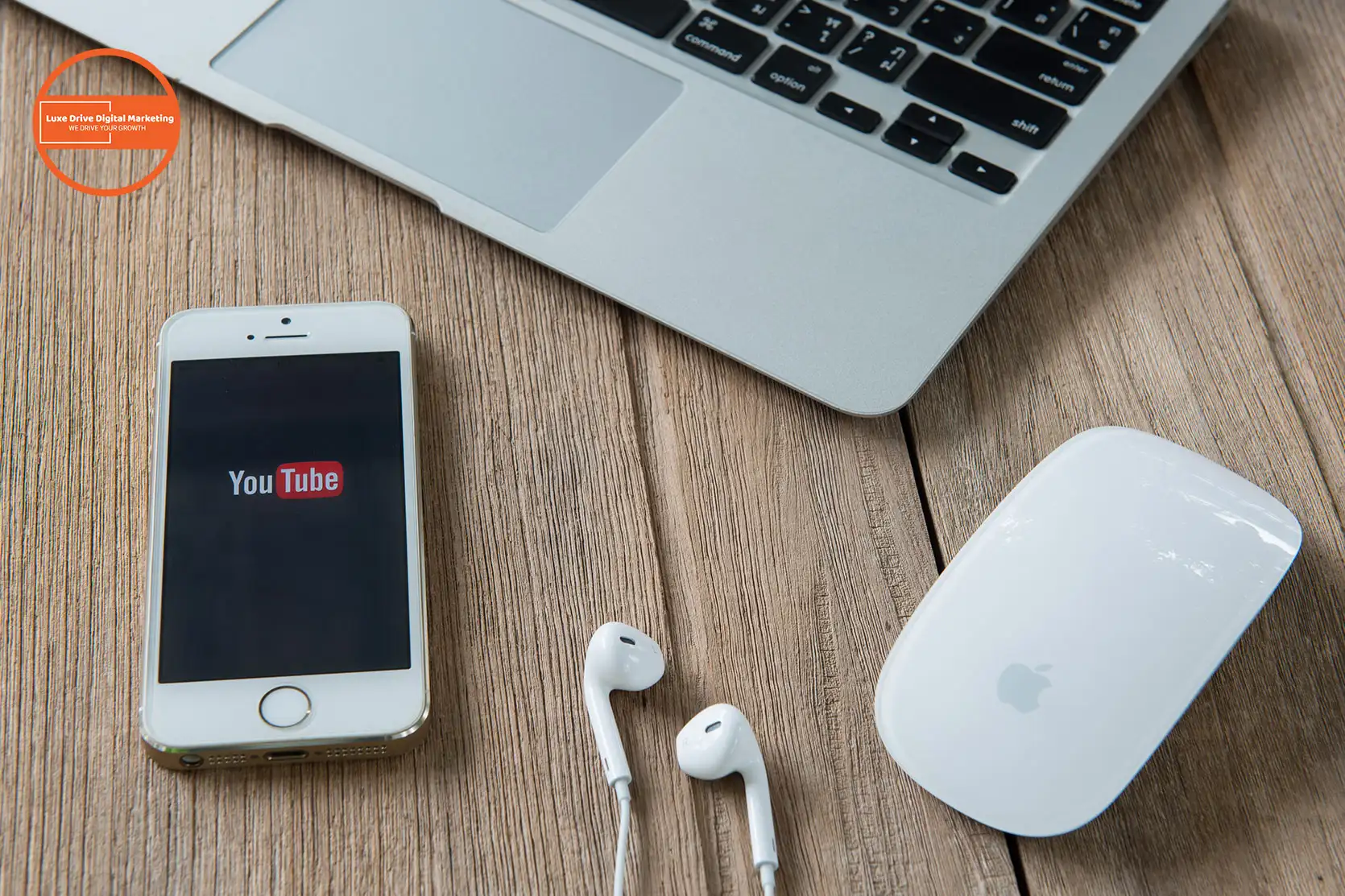 Luxe Drive Digital Marketing America's Top Marketing Agency An iPhone displaying the YouTube app, white earphones, a white Apple Magic Mouse, and a laptop are perfectly arranged on a Luxe Drive wooden surface. Luxe Drive Digital Marketing We Drive Your Growth