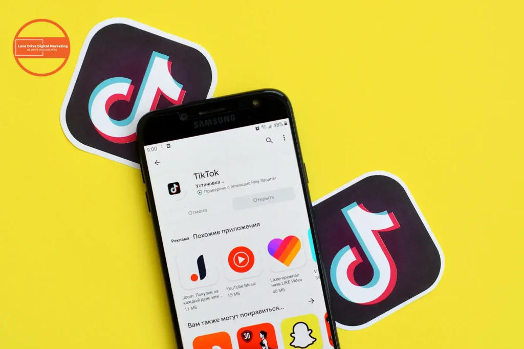 Luxe Drive Digital Marketing America's Top Marketing Agency A smartphone displaying the TikTok app page on its screen, placed on a yellow surface with two TikTok logo stickers nearby, evokes the essence of Luxe Drive digital marketing. Luxe Drive Digital Marketing We Drive Your Growth