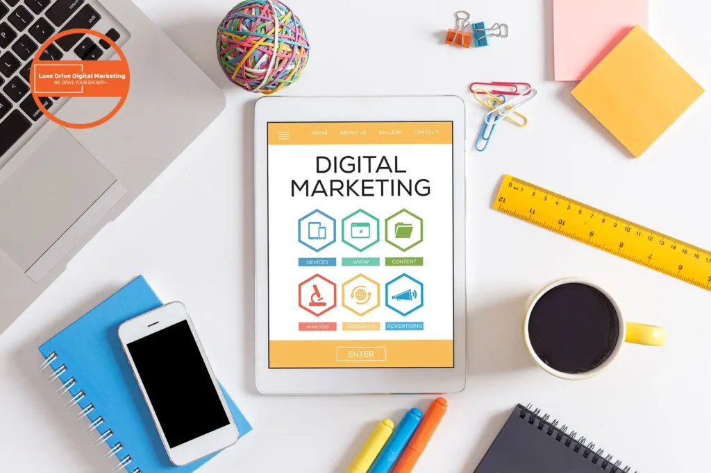 Luxe Drive Digital Marketing America's Top Marketing Agency Tablet displaying "Digital Marketing" surrounded by a laptop, smartphone, coffee cup, colorful pens, ruler, paper clips, and a rubber band ball on a white desk. Luxe Drive Digital Marketing We Drive Your Growth