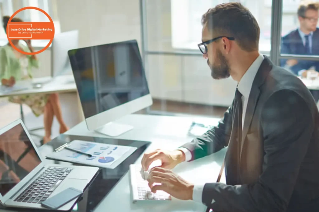 Luxe Drive Digital Marketing America's Top Marketing Agency Man in a suit working on a computer at a desk, with charts on the screen. A laptop is open beside him. Another person is blurred in the background. Circular Luxe Drive Digital Marketing logo. Luxe Drive Digital Marketing We Drive Your Growth