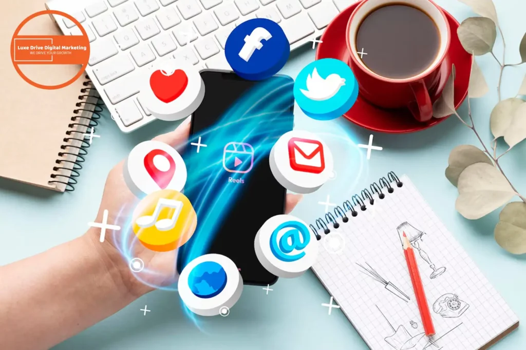 Luxe Drive Digital Marketing America's Top Marketing Agency A hand holding a smartphone with social media icons floating around. Nearby are a red cup, a notebook with a doodle, and a branch with leaves on a light surface. Luxe Drive Digital Marketing We Drive Your Growth