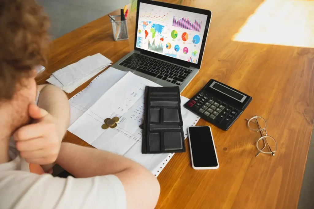 Luxe Drive Digital Marketing America's Top Marketing Agency Person analyzing charts on a laptop with papers, coins, a calculator, phone, and glasses on a wooden table. Luxe Drive Digital Marketing We Drive Your Growth