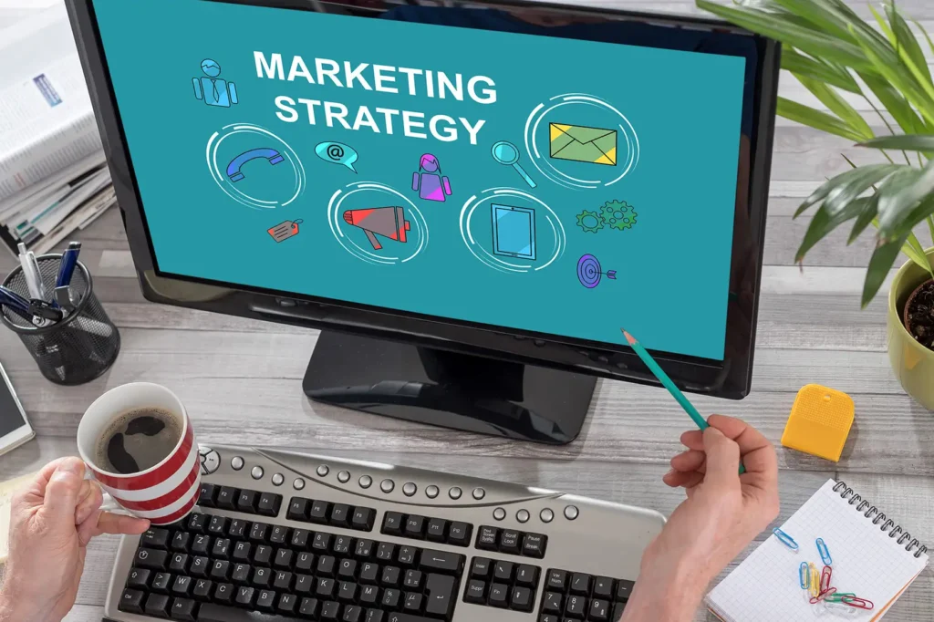 Luxe Drive Digital Marketing America's Top Marketing Agency Computer screen displaying "Marketing Strategy" with related icons. Person points with a pencil while holding a coffee cup, seated at a desk with a keyboard, notebook, and office supplies. Luxe Drive Digital Marketing We Drive Your Growth